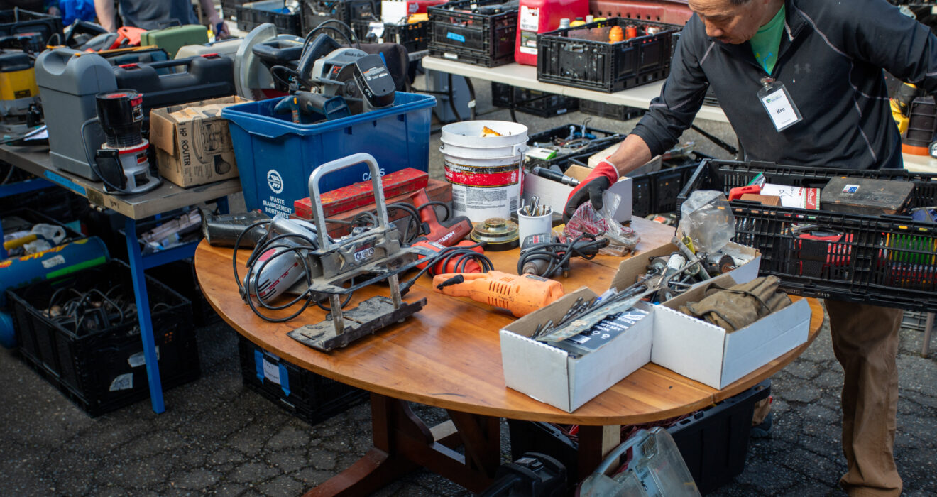 An individual holding some tools while taking a look at the abundance of tools on the table at the Shoreline Tool Sale. There are plenty of tools organized on tables all around them.