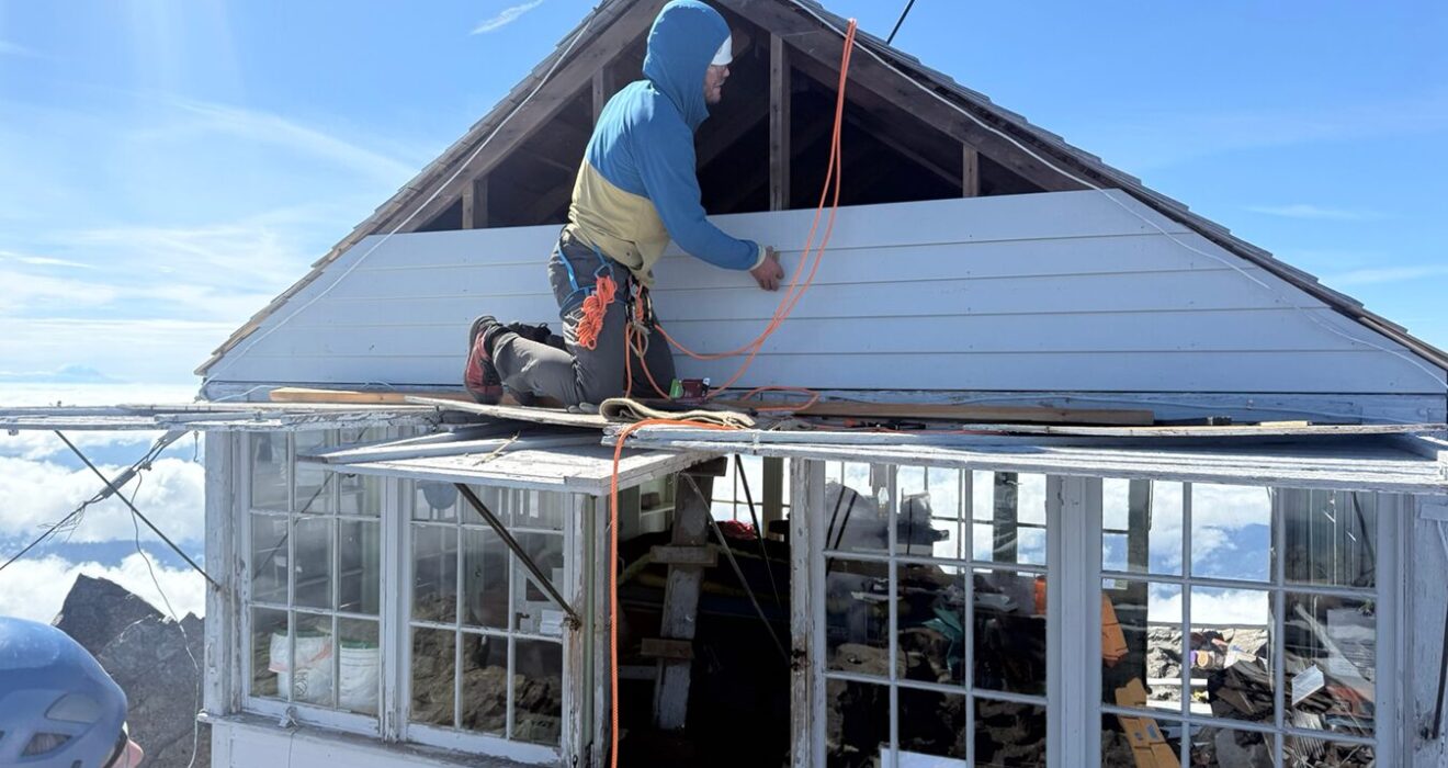 A volunteer sits on top of the roof of Three Fingers Lookout while someone nearby observes.