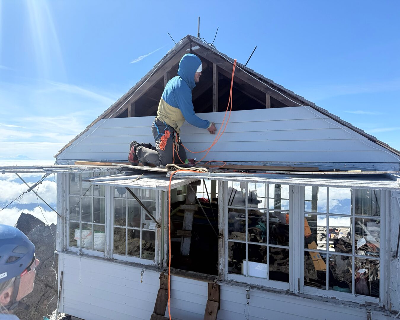 A volunteer sits on top of the roof of Three Fingers Lookout while someone nearby observes.