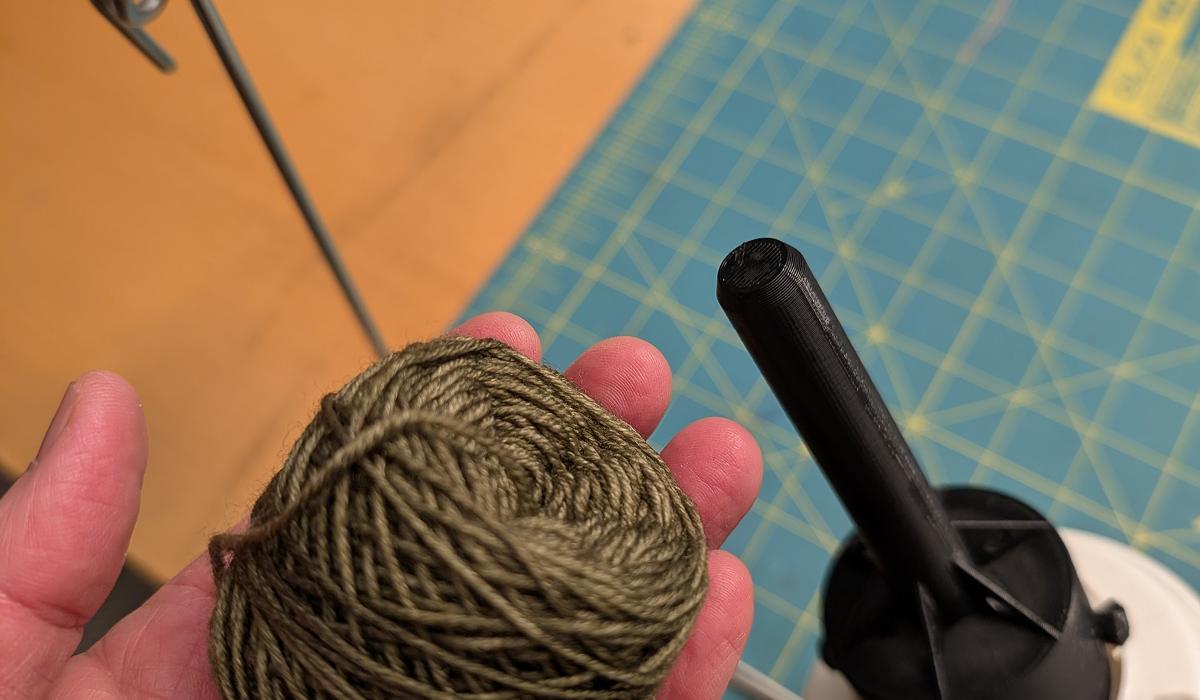 A hand holds a small skein of yarn that was created with the 3D printed yarn winder adaptor in the background.