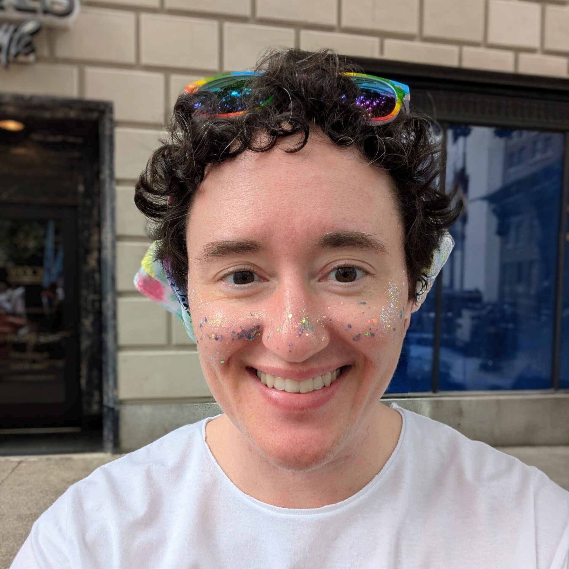 A selfie of Kahle Gorzelsky, wearing colorful facial glitter, sunglasses on their head, and a head scarf.