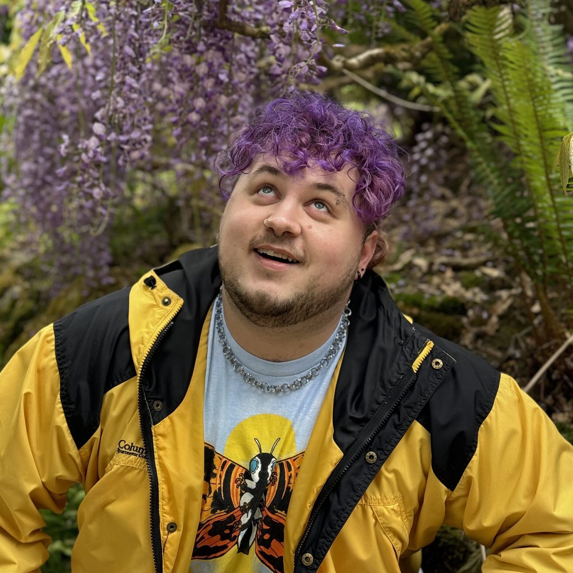 With purple hair and a yellow jacket, Gage smiles up at the purple plant above him.