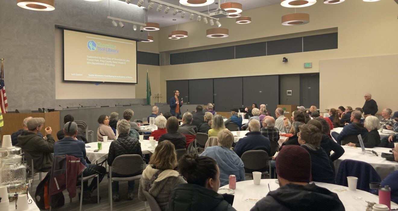 Someone gives a presentation at the front of a room full of people sitting at different tables.