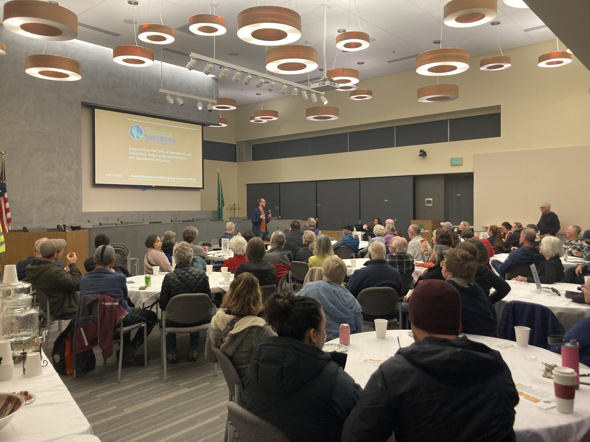 Someone gives a presentation at the front of a room full of people sitting at different tables.